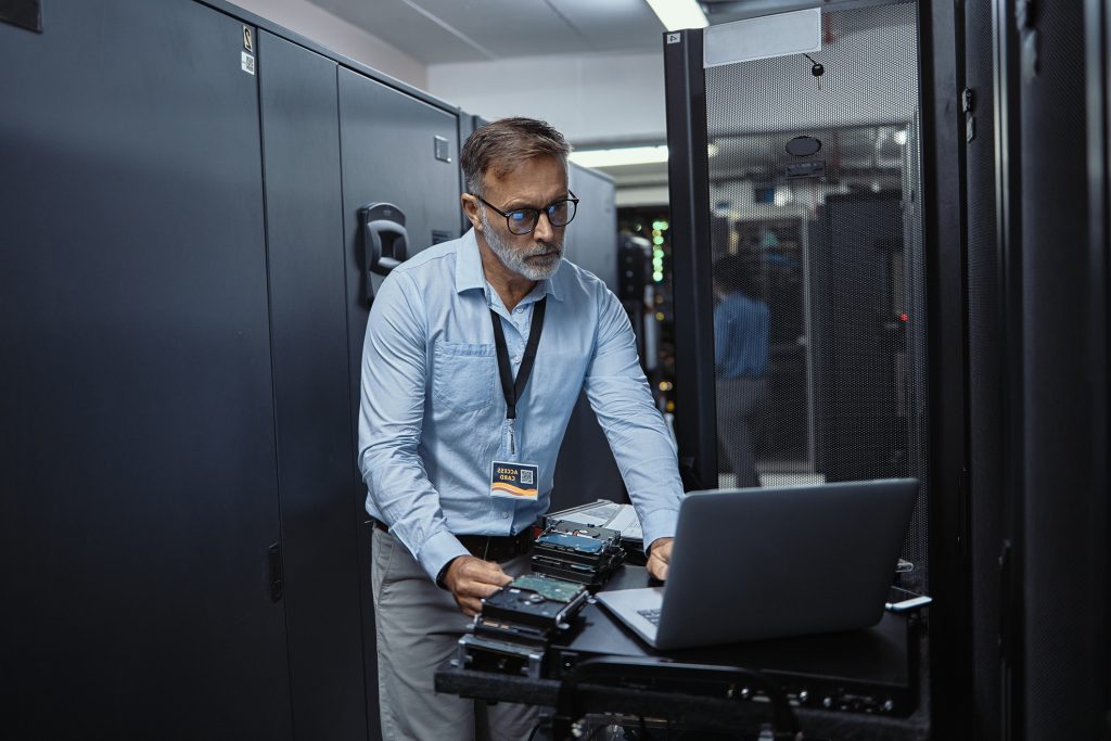 Mature software engineer using a laptop in a server room. IT technician working on network programming with online technology. Protecting information on the internet with cyber security management